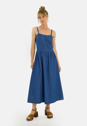 Femme aux cheveux blonds en chignon portant une robe midi en denim bleu sans manches et des sandales compensées marron, debout devant un fond uni.