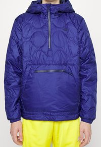 Marinblå, vadderad pulloverjacka med huva, med halvdragkedja framtill, ficka med dragkedja och elastiska handledar. Bärs över gula byxor.