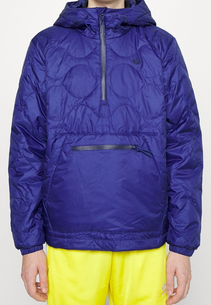 Marinblå, vadderad pulloverjacka med huva, med halvdragkedja framtill, ficka med dragkedja och elastiska handledar. Bärs över gula byxor.