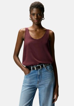 Donna con i capelli corti che indossa orecchini a cerchio, canotta bordeaux, jeans blu e cintura nera, in piedi con una mano in tasca.