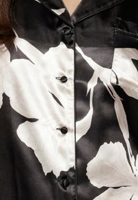 Zwarte satijnen blouse met grote witte bloemenpatronen, zes zwarte knopen en een opstaande kraag. Glad materiaal en op maat gemaakte pasvorm.