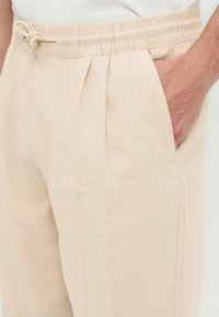Pantalon en lin beige avec taille élastique et cordon de serrage, une main insérée dans la poche latérale droite, porté avec un haut blanc.