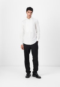 Witte dressshirt met verticale plooien aan de voorkant, lange mouwen en een ronde zoom, gecombineerd met zwarte broek en zwarte schoenen.
