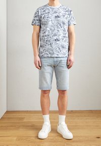 White t-shirt with blue leaf pattern, paired with light blue denim shorts. White sneakers and crew-length socks complete the outfit.