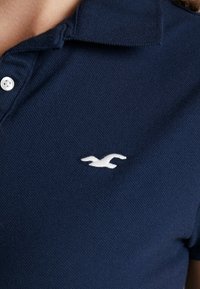 Marinblå pikétröja i texturerad tyg, med en vit broderad logotyp på bröstet och en treknappskrage.