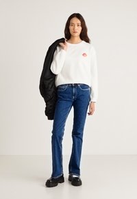 Sudadera blanca con un gráfico de labios rojos, vaqueros azul oscuro y botas negras con plataforma. La modelo sostiene una chaqueta negra. Estilo simple y casual.