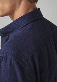 Persoon in een donkerblauw overhemd met kraag, gezien vanaf de achterkant, waarbij het schouder- en nekgebied zichtbaar is.