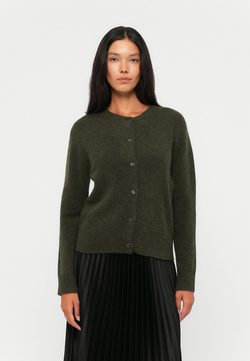 Textured dark green cardigan with a round neckline, front button closure, and long sleeves, paired with a pleated black skirt.