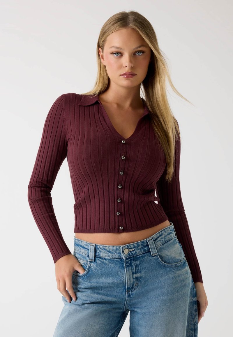 Bordeauxroter gerippter Langarmstrickcardigan mit V-Ausschnitt und Knopfverschluss. Figurbetontes Design, weiche Textur, kombiniert mit hellblauen Jeans.