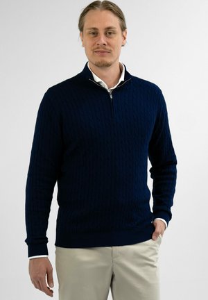 Mann mit hellbraunen Haaren, der einen marineblauen, strukturierten Pullover mit Viertelreißverschluss und beige Hosen trägt, steht vor einem schlichten weißen Hintergrund.