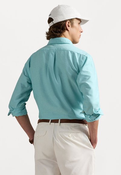 Gaiši zils krekls ar rolled piedurknēm, izgatavots no gludas auduma. Nēsāts ar bežīgiem biksēm un baltu cepuri. Redzams brūns josta.