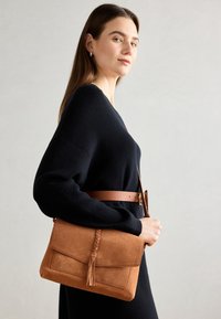 Suede skuldertaske i tan med en flettet detalje og frynse. Har en frontklap og en brun læderrem.