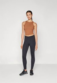 Barna ujjatlan rövid felső, magas derekú fekete leggingsszel párosítva. A modell fekete, texturált részletekkel ellátott sportcipőt visel.