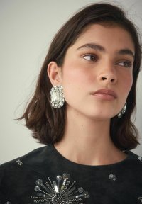 Junge Frau mit kurzen braunen Haaren, die große rechteckige Kristallohrringe und ein schwarzes Oberteil mit silbernen Pailletten trägt.