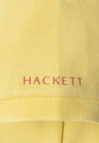 Żółty bawełniany sweter z prążkowanymi mankietami, z wyraźnym czerwonym logo "HACKETT" na dole. Tekstura jest gładka i dziana.