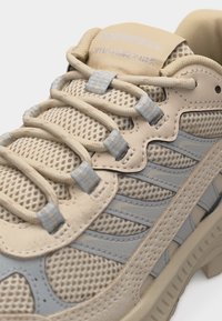 Μπεζ και γκρι πεζοπορικό παπούτσι Merrell που δείχνει το επάνω μέρος από δίχτυ, τη μαλακή γλώσσα και τα ασφαλή κορδόνια περασμένα μέσα από υφασμάτινους βρόχους.
