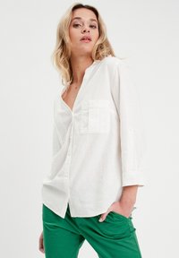 Chemise en lin blanche avec un col, poche avant et fermeture à boutons. Portée avec un pantalon vert, montrant une coupe décontractée et une texture douce.