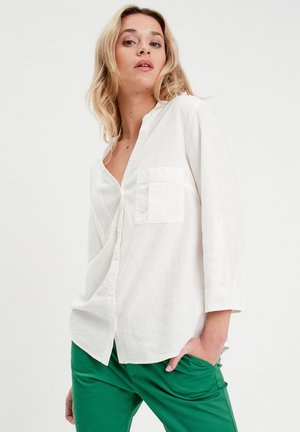 Camisa de lino blanca con cuello, bolsillo frontal y cierre de botones. Llevada con pantalones verdes, que muestran un corte relajado y una textura suave.