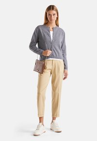 Šedý kardigan s kulatým výstřihem a zapínáním na knoflíky, spárovaný s béžovými kalhotami a bílými teniskami; modelka drží malou hnědou tašku.