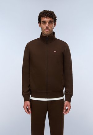 Uomo con capelli ricci che indossa una giacca marrone con zip e pantaloni abbinati, in piedi davanti a uno sfondo chiaro uniforme.