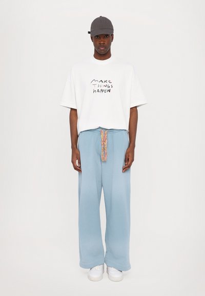 Hvid oversized t-shirt med teksten "Gør Ting Til Virkelighed", parret med lysblå bredbenede bukser med en multifarvet snor, båret med hvide sneakers.