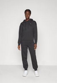 Frau, die in einem schwarzen Puma-Hoodie und passenden Jogginghosen mit weißen Sneakers vor einem schlichten hellen Hintergrund steht.