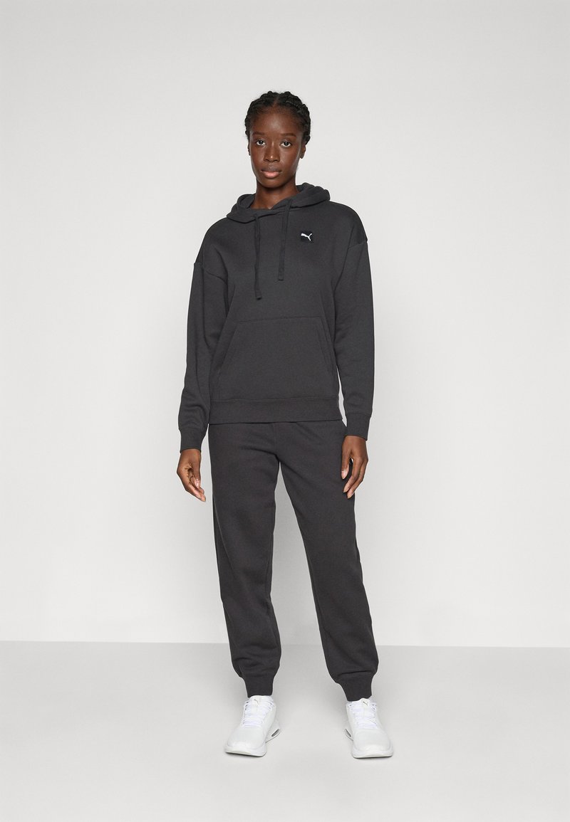 Frau, die in einem schwarzen Puma-Hoodie und passenden Jogginghosen mit weißen Sneakers vor einem schlichten hellen Hintergrund steht.