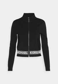 Giacca nera con zip e colletto alto, caratterizzata da una fascia bianca a contrasto all'orlo con testo "MICHAEL KORS". Texture liscia, design aderente.