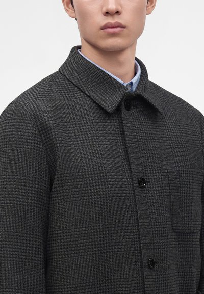 Jeune homme portant un manteau en laine à carreaux gris foncé avec col et boutons noirs par-dessus une chemise bleu clair.