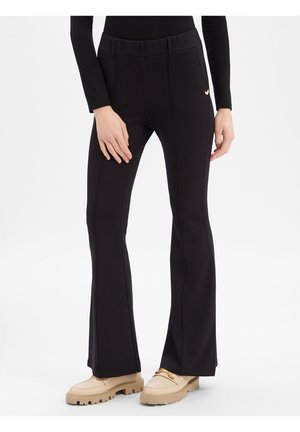 Femme portant un pantalon flare taille haute noir avec des coutures verticales, associé à des mocassins beige épais et un haut noir à manches longues.