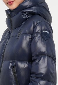 Tamsiai mėlynas pūkinis švarkas su blizgiu paviršiumi, aukšta apykakle ir siuvinėjimo detalėmis. Yra šoninis užtrauktukas kišenė ir pleistras ant kairiojo rankovės.