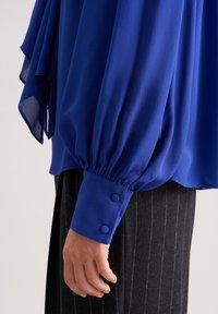 Blusa blu royal in tessuto strutturato, con maniche a sbuffo e polsini aderenti con due bottoni coordinati. Indossata con pantaloni a righe.