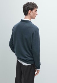 Tmavě modrý pletený svetr s límcem, dlouhými rukávy a měkkou texturou; nošený přes bílou košili s límcem, doplněný o tmavé kalhoty.