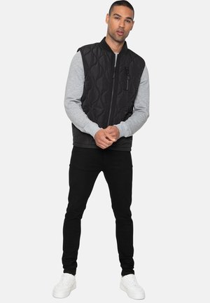 Black quilted vest with a gray long-sleeve base, front zip closure, and side pockets, paired with black pants and white sneakers.
