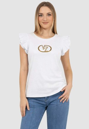 Giovane donna che indossa una maglietta bianca con maniche arricciate e logo YZ dorato, insieme a jeans blu, in piedi davanti a uno sfondo grigio chiaro.