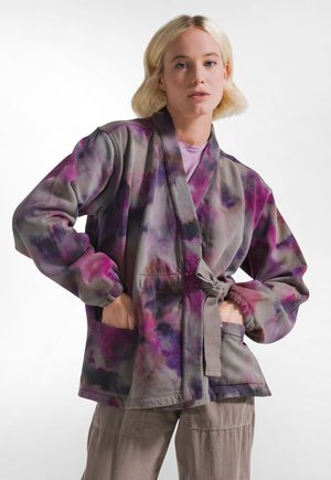 Donna bionda che indossa una giacca larga tie-dye nei toni del grigio, viola e rosa, con le mani nelle tasche ed espressione neutra.