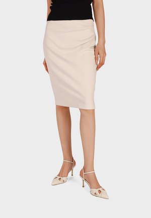 Donna che indossa una gonna a matita beige al ginocchio, top nero senza maniche e scarpe beige con tacchi alti e cinturini, in piedi su uno sfondo grigio.