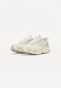 Chaussures de trail running légères beiges claires avec une tige en mesh, une semelle texturée, un motif de crampons prononcé et un laçage avec des accents pastel.