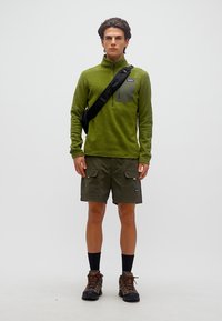 Νέος άνδρας φοράει φλις πουλόβερ ελιάς, σορτς cargo, μαύρη τσάντα χιαστή, μαύρες κάλτσες και ορειβατικά μποτάκια, στέκεται μπροστά σε λευκό φόντο.