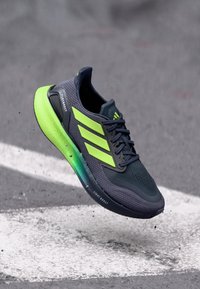 Chaussure de running Adidas avec une tige en mesh gris foncé, des accents verts vifs, et une semelle rembourrée et texturée au design dégradé.