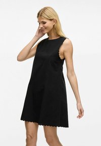 Robe noire sans manches avec des bordures festonnées, fabriquée dans un tissu léger, ayant une coupe ample et un design épuré et minimaliste.
