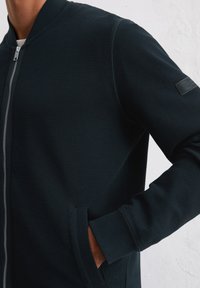 Dark blue textured bomber jacket with a front zipper, ribbed cuffs, and side pockets. Features a subtle logo patch on the sleeve.
