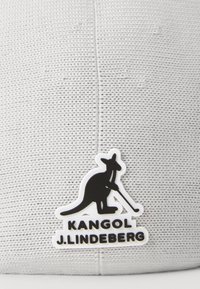 Căciulă gri țesută cu o suprafață texturată, având un logo de cangur negru și textul "KANGOL J.LINDEBERG" în litere albe îngroșate.