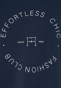 Námornícka látka s bielym textom usporiadaným do kruhu s nápisom „EFFORTLESS CHIC FASHION CLUB“ a štýlovo upravené logo „FM“ s nápisom „SINCE 1968“ vo vnútri.