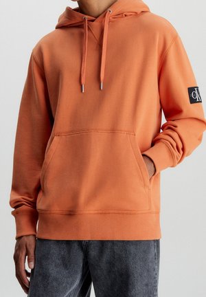 Muž nosící oranžovou mikinu s kapucí a přední kapsou a černou nášivkou s logem Calvin Klein na horním rukávu.