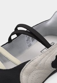 Povečani posnetek črno-belega čevlja z izrezljanim velurjem in dvema črnima elastičnima trakoma čez zgornji del.