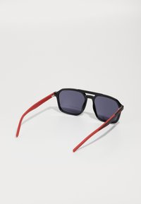 Ochelari de soare negri cu un cadru geometric, lentile închise la culoare și sprijine din cauciuc roșu, având o textură elegantă și un design unghiular.
