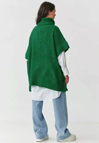 Kobieta w grubej, zielonej ponczo z golfem, nosząca długą białą koszulę i jasnoniebieskie spodnie typu wide-leg, stoi odwrócona plecami.