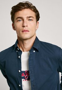 Camisa azul marino de botones en tejido ligero con un acabado texturizado, que presenta un logo sutil y un cuello, superpuesta a una camiseta gráfica blanca.