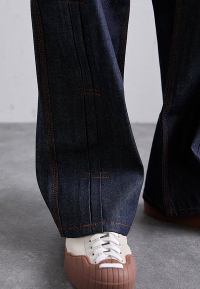 Wide dark blue denim pants with orange stitching, paired with white and brown sneakers featuring a textured rubber sole.